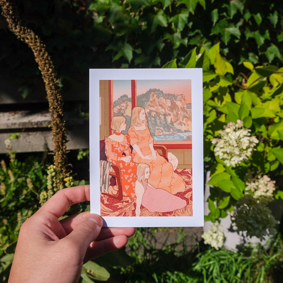 Hand holding a postcard with a colorful illustration of three women in a room, surrounded by greenery.